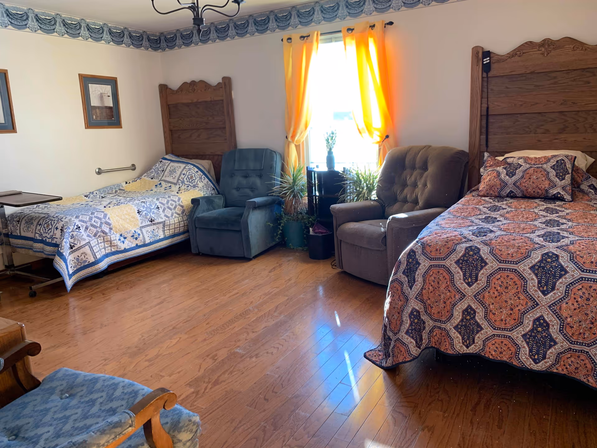 A cozy bedroom with two beds featuring wooden headboards and patterned quilts. Between the beds are two armchairs and a small table with plants in front of a window with yellow curtains. The room has wooden flooring and framed pictures on the walls.
