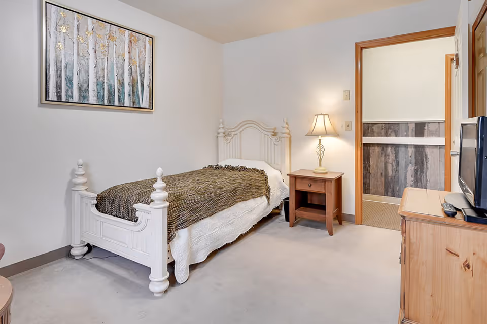 A small bedroom with a white wooden single bed covered with a white bedspread and a brown knitted blanket. Next to the bed is a wooden nightstand with a lamp on it. A wooden dresser with a TV and remote controls is on the right side. The walls are white, and there is a framed painting of trees hanging above the bed. An open doorway leads to another room with wood-paneled walls.