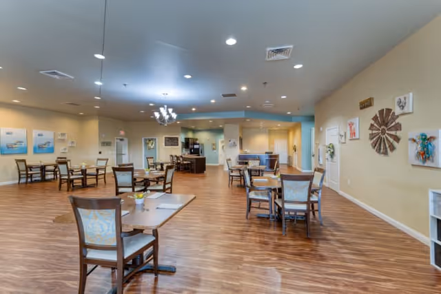 A spacious dining room with multiple wooden tables and chairs arranged neatly on a wooden floor. The walls are decorated with artwork and a decorative windmill piece. The ceiling has recessed lighting and a chandelier. In the background, there is a kitchen or serving area with counters and appliances.