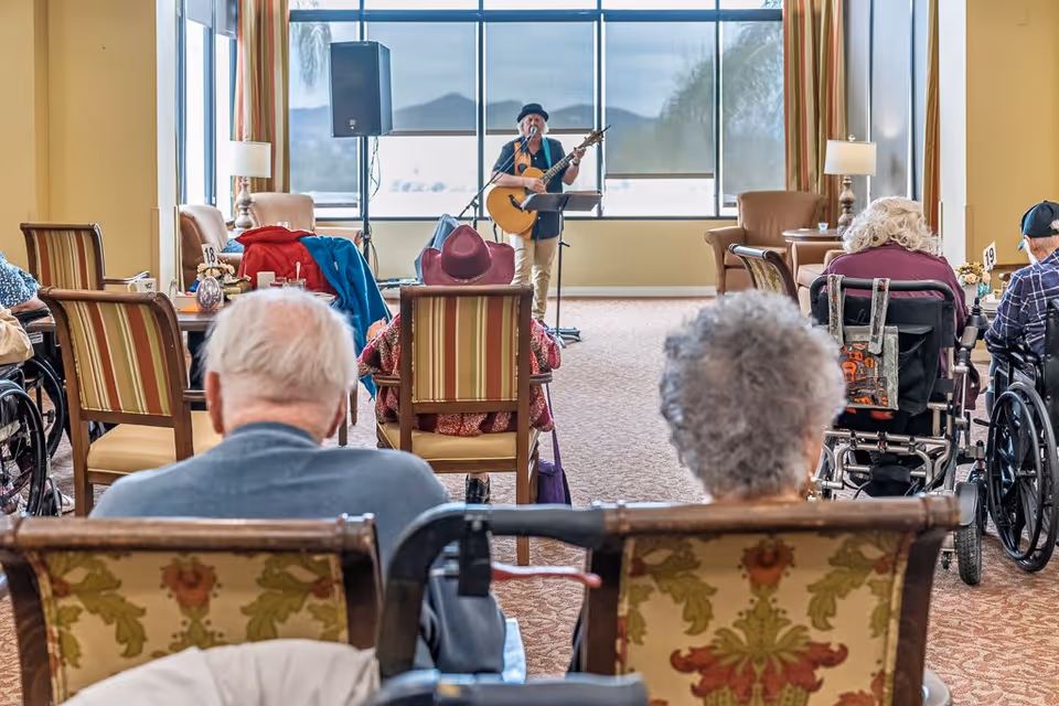 A group of elderly people seated in a common area of a senior living facility, watching a musician playing guitar and singing in front of large windows.