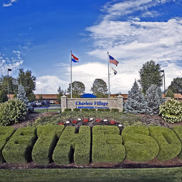 Outdoor view of the entrance sign for Charless Village, a Bethesda community, with neatly trimmed bushes spelling out 'BETHESDA' in front. Three flagpoles with flags are visible behind the sign, along with trees and a blue sky with clouds.