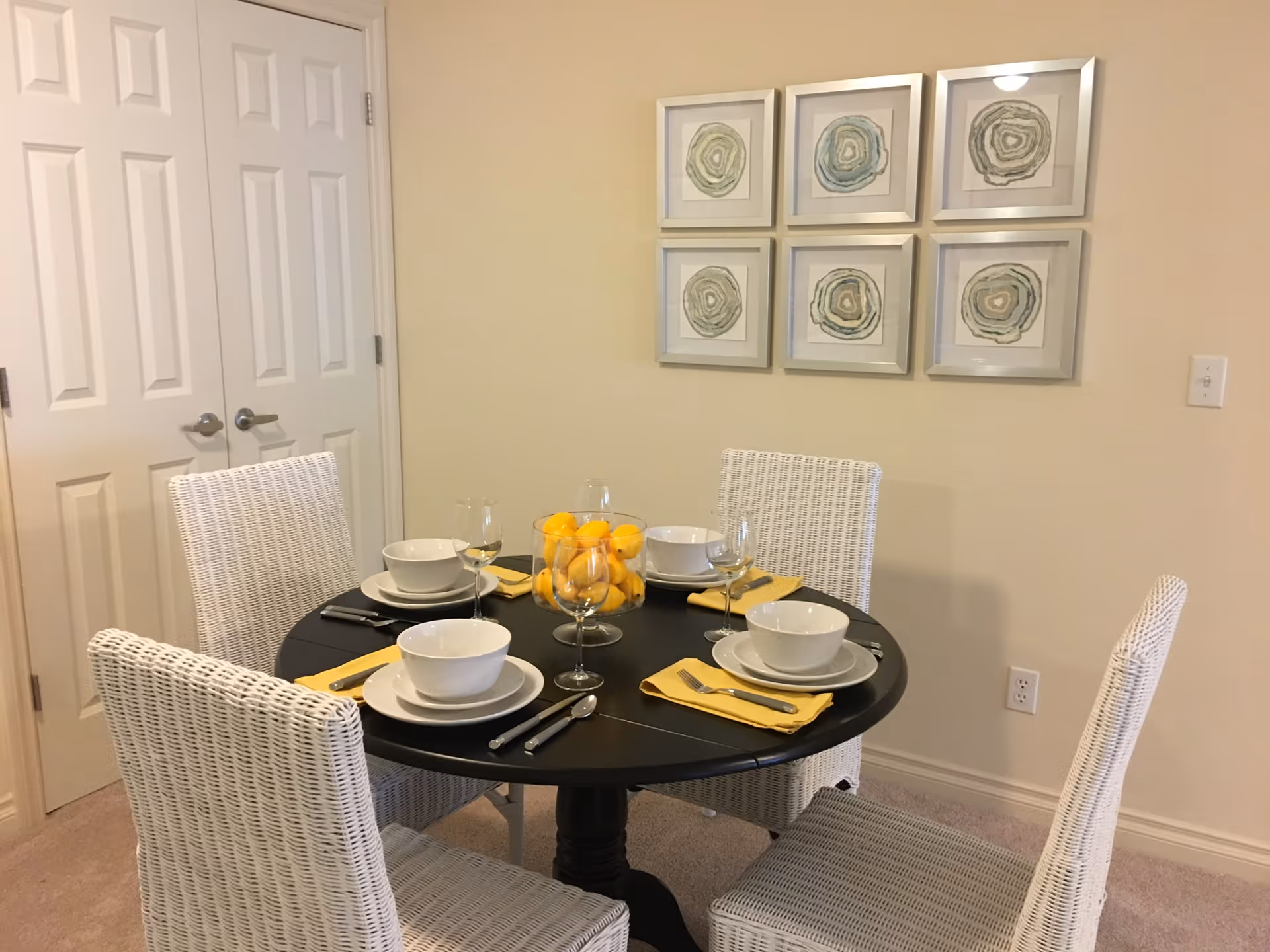 A dining area with a round black table set for four people. Each place setting includes a white bowl, plate, wine glass, and yellow napkin with silverware. The chairs are white wicker. On the table is a glass bowl filled with yellow lemons. The background shows a beige wall with six framed abstract art pieces and a double white door.