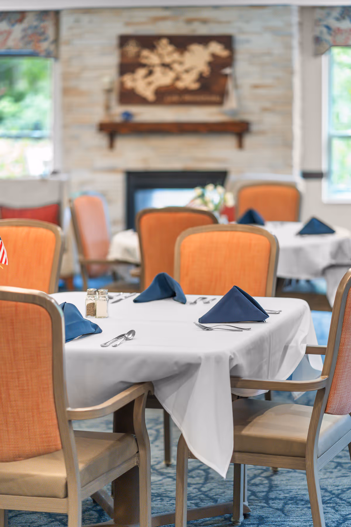 A dining room with tables covered in white tablecloths, each set with blue folded napkins, silverware, and salt and pepper shakers. The chairs have orange upholstery and wooden frames. In the background, there is a stone fireplace with a wooden mantel and a decorative wall piece above it. Windows on either side let in natural light.
