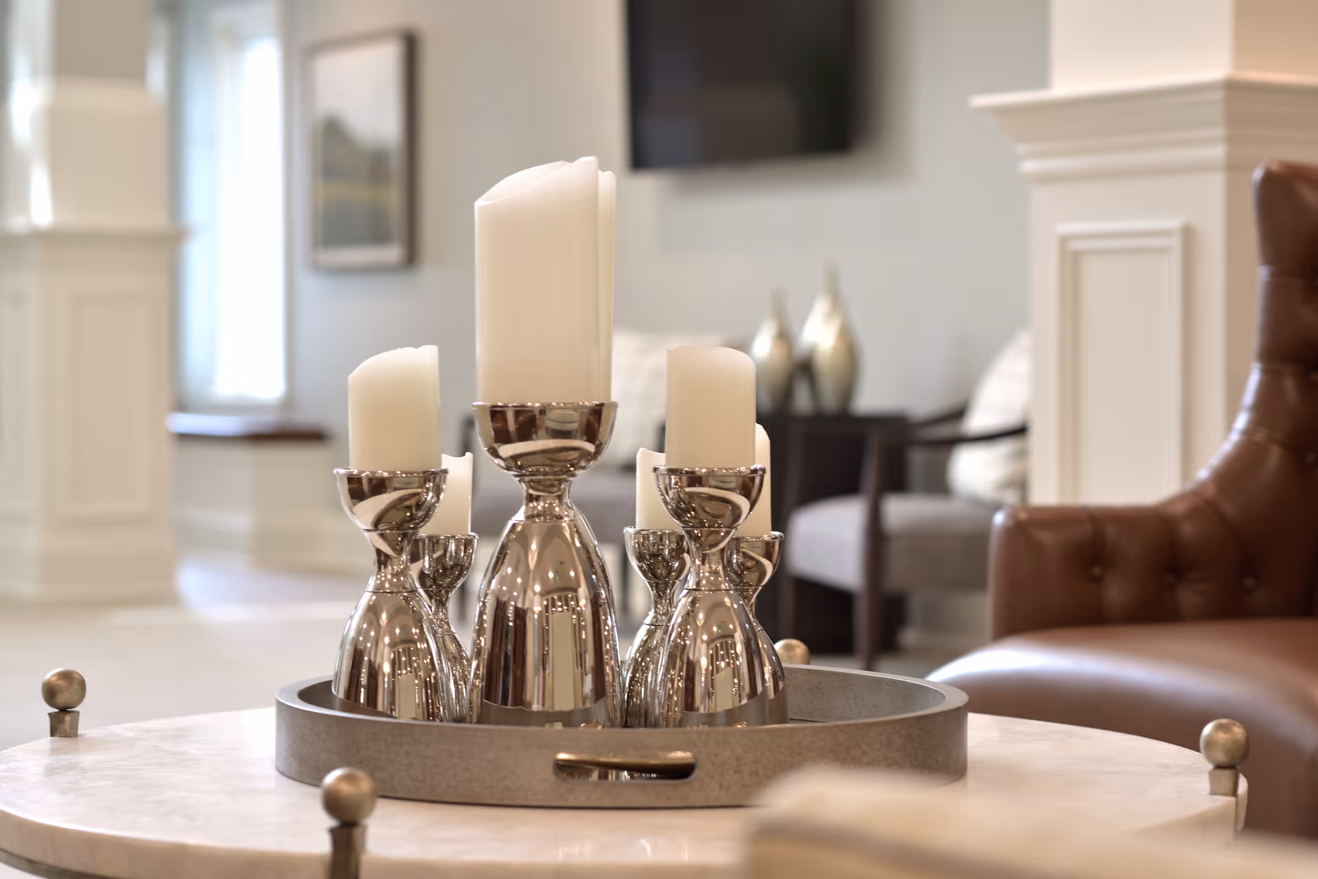 Close-up of a decorative tray with silver candle holders and white candles on a marble table in a cozy living room setting with a brown leather chair and soft lighting.