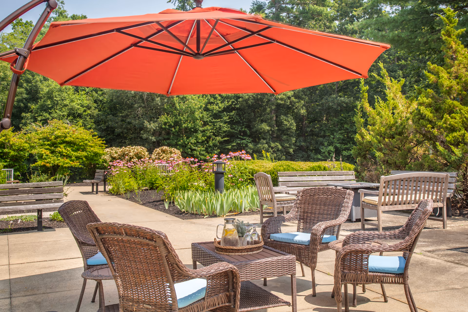 Outdoor patio with wicker chairs and a table under a large red umbrella overlooking garden beds and benches.
