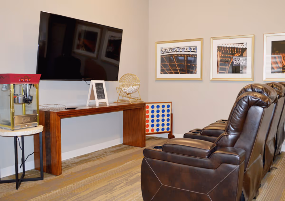 A cozy entertainment room with four brown leather recliner chairs facing a large flat-screen TV mounted on a beige wall. Below the TV is a wooden console table holding a bingo cage and a small chalkboard sign. To the left, there is a popcorn machine on a small round table. On the wall behind the chairs are three framed pictures with theater-themed images. A large Connect Four game is positioned against the wall near the console table.
