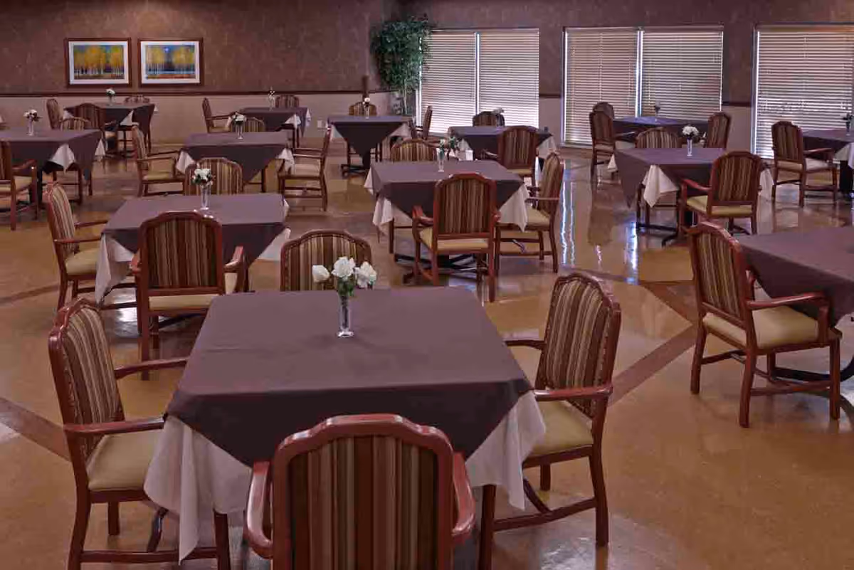 A dining room with multiple square tables covered with white and dark brown tablecloths, each table surrounded by wooden chairs with striped cushions. Small vases with white flowers are placed on each table. The room has large windows with blinds and framed artwork on the walls.