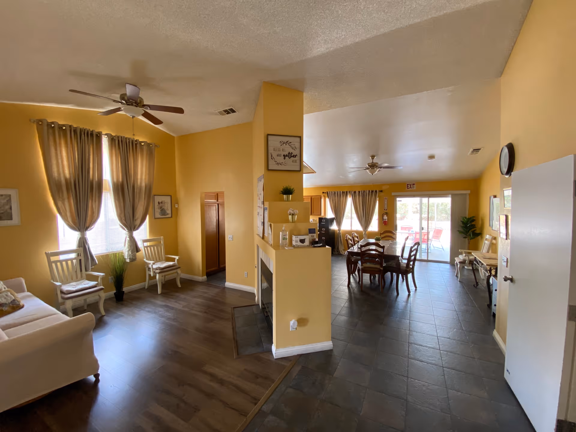 Interior view of a senior living facility common area with yellow walls and two ceiling fans. The space is divided into a sitting area with a white couch, two chairs, and a window with curtains on the left, and a dining area with a wooden dining table and chairs on the right. There is a fireplace in the center wall, decorative plants, framed pictures, and a sliding glass door leading to an outdoor patio.