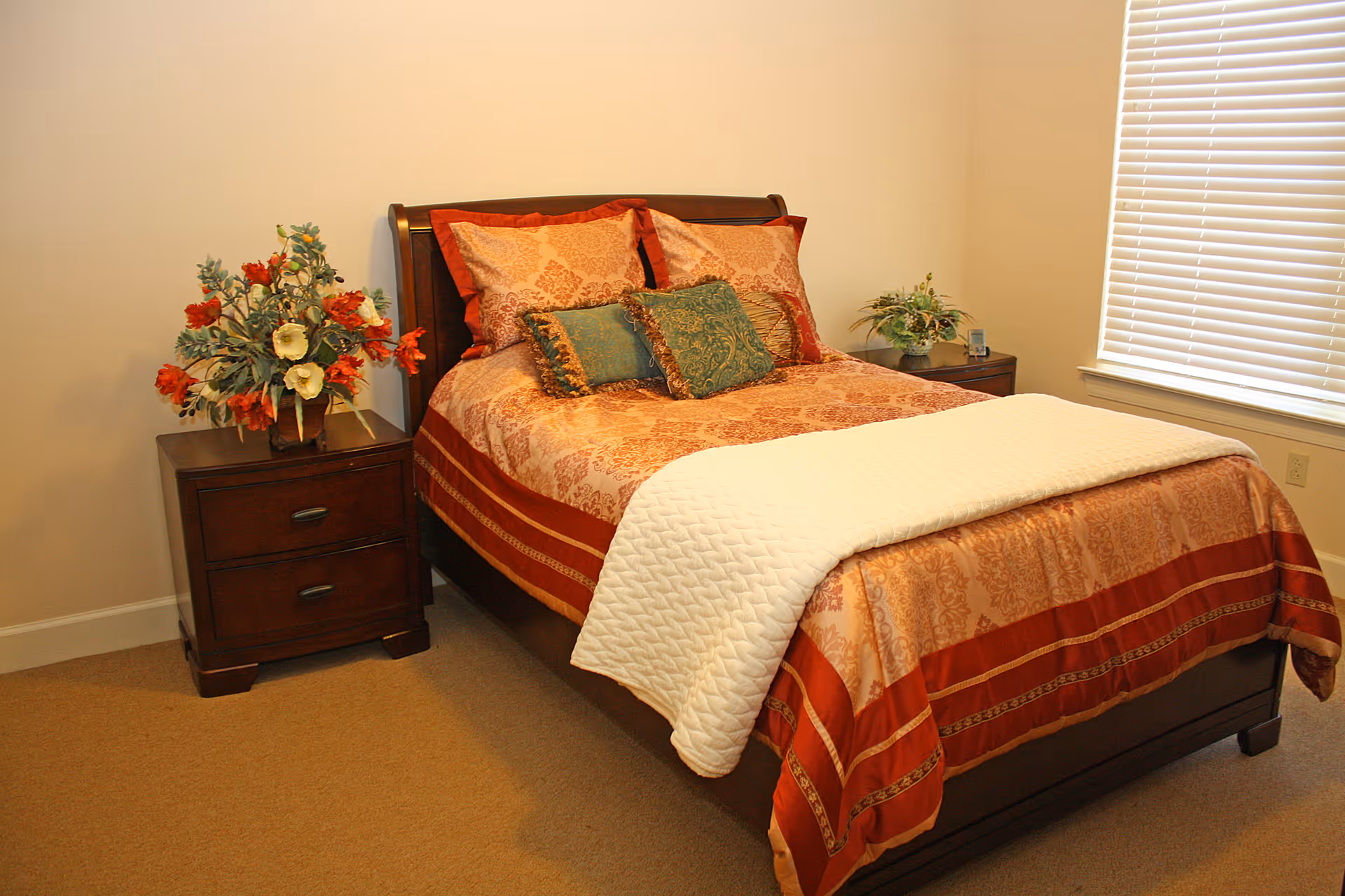 A neatly made bed with a patterned red and gold bedspread and decorative pillows in a bedroom. There are two wooden nightstands on either side of the bed, each with a floral arrangement. A window with closed blinds is visible on the right side of the room.