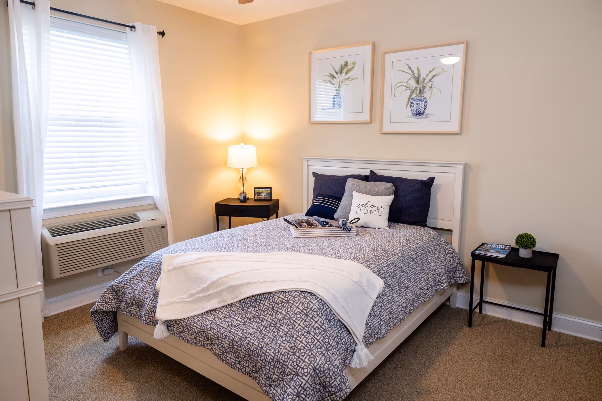 A cozy bedroom with a neatly made bed featuring a blue patterned bedspread, multiple pillows including one with the text 'welcome HOME', and a white throw blanket. There are two framed botanical prints hanging on the beige wall above the bed. A window with white blinds and sheer curtains is on the left side, with an air conditioning unit below it. Two black side tables flank the bed, one with a lamp and a small framed photo, and the other with a small green plant and a magazine.