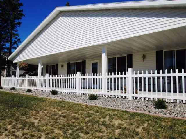 Exterior view of a single-story building with white siding and a covered porch area. The porch is enclosed by a white picket fence, and there are several windows and doors along the building's facade. The ground in front of the fence has rocks and small shrubs, with a grassy area in the foreground.