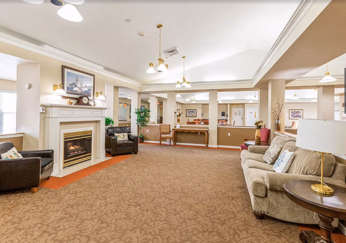 Spacious senior living common area with beige carpet, a fireplace with a clock and framed picture above it, two black armchairs with decorative pillows, a beige sofa with patterned pillows, a round wooden side table with a lamp, and multiple ceiling light fixtures. The room has beige walls with white trim and several columns, with a dining area visible in the background.