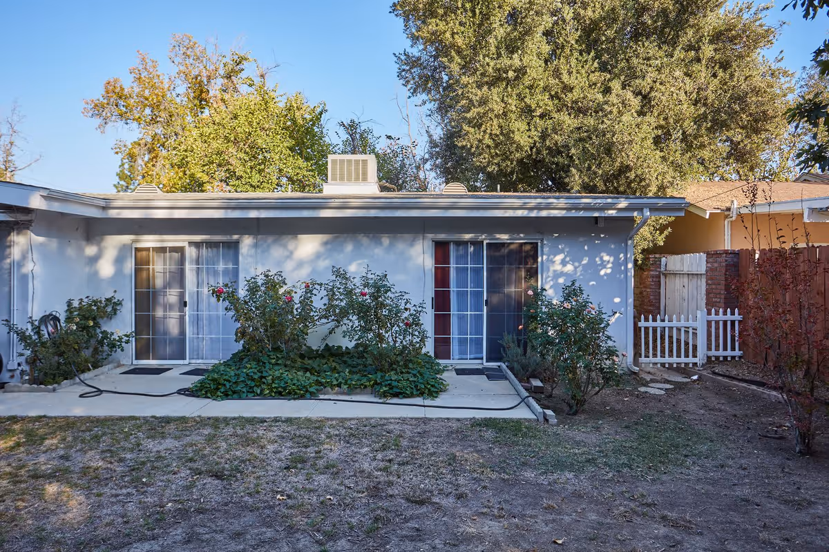 Exterior view of a single-story building with two sliding glass doors, surrounded by plants and bushes. There is a small concrete patio in front of the doors and a white picket gate to the right. Trees and a clear blue sky are visible in the background.