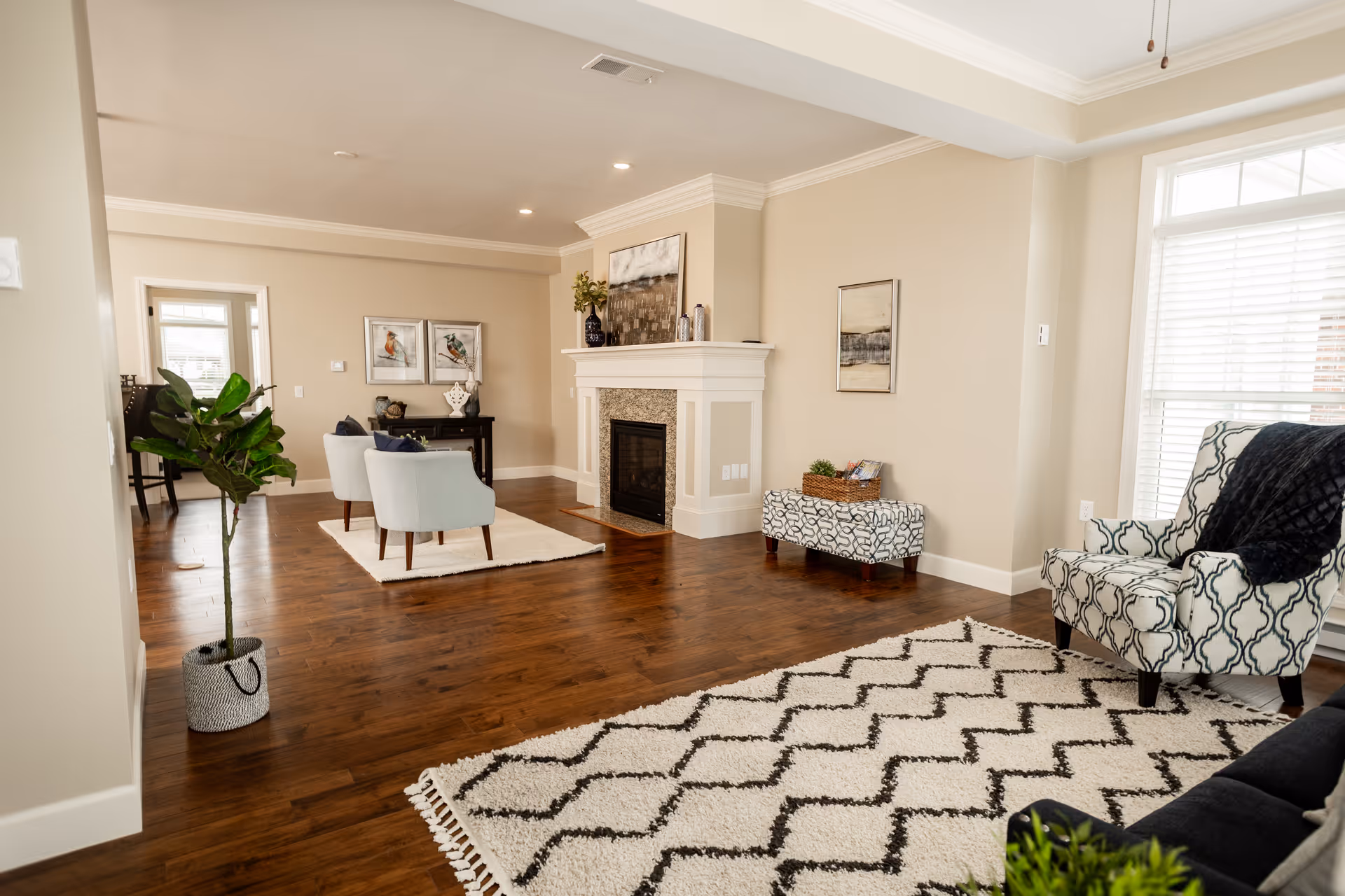 A bright and spacious living room with wooden floors, a white fireplace, and large windows with blinds. The room features patterned armchairs, a black sofa, a white and black geometric rug, a small ottoman with a basket on top, and a potted plant. There are framed artworks on the walls and recessed ceiling lights.
