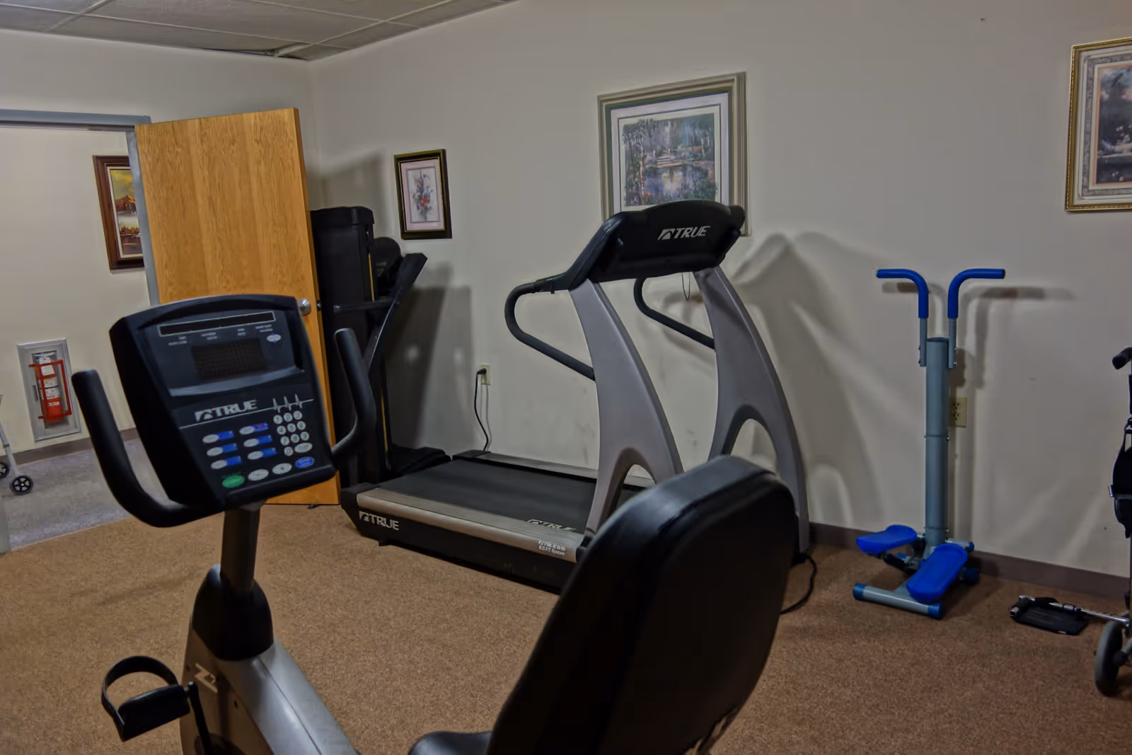 Small fitness room with a treadmill, stationary bike, and a blue stepper against a beige wall with framed pictures.