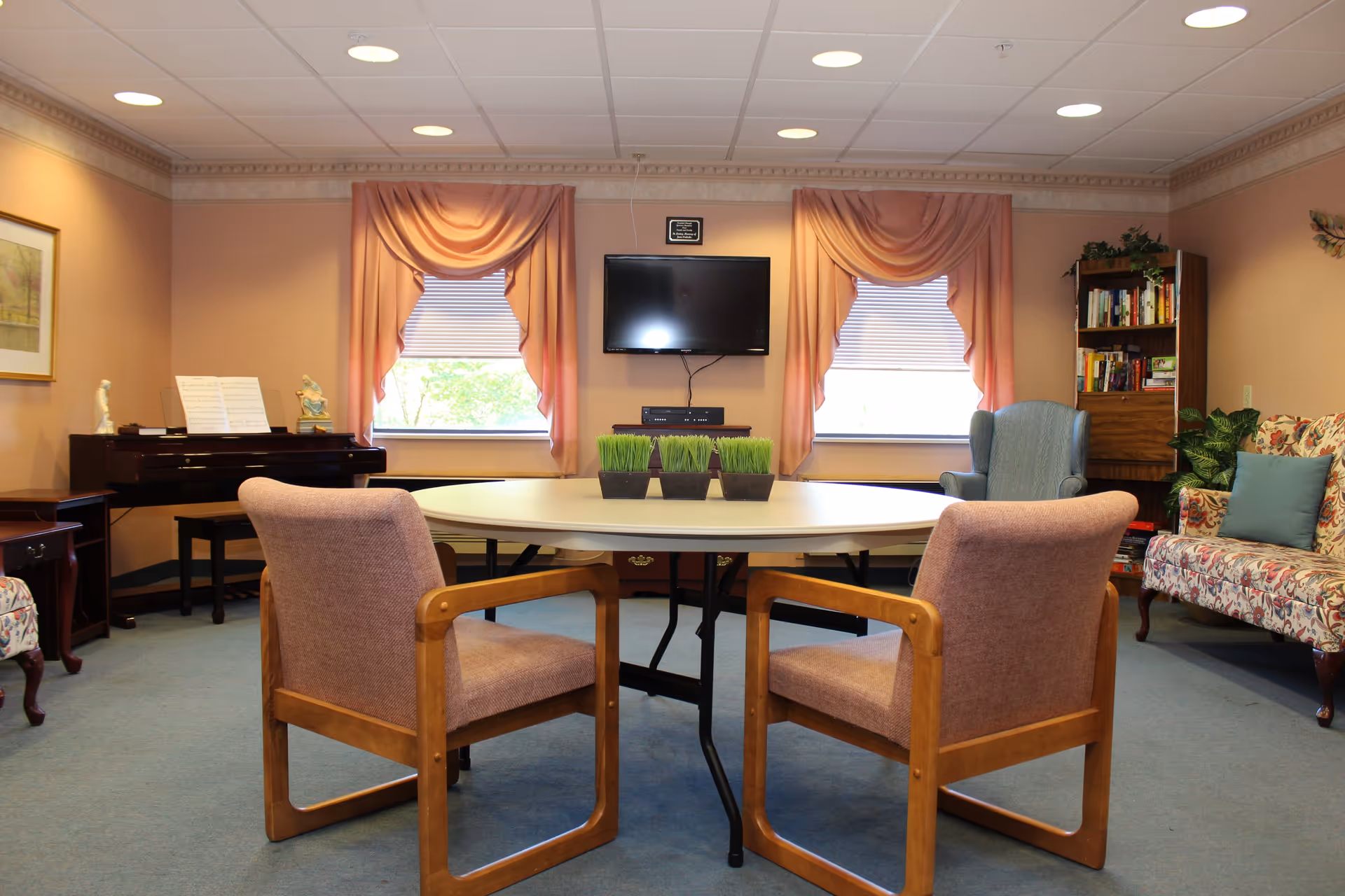 A cozy common room with a round table and two wooden armchairs in the center. Behind the table are two windows with pink drapes, a wall-mounted flat-screen TV, and a small cabinet. To the left, there is a piano with sheet music and decorative figurines. On the right side, there is a blue armchair, a floral-patterned sofa with a blue pillow, a bookshelf filled with books, and some green plants. The room has soft peach-colored walls and a blue carpeted floor.