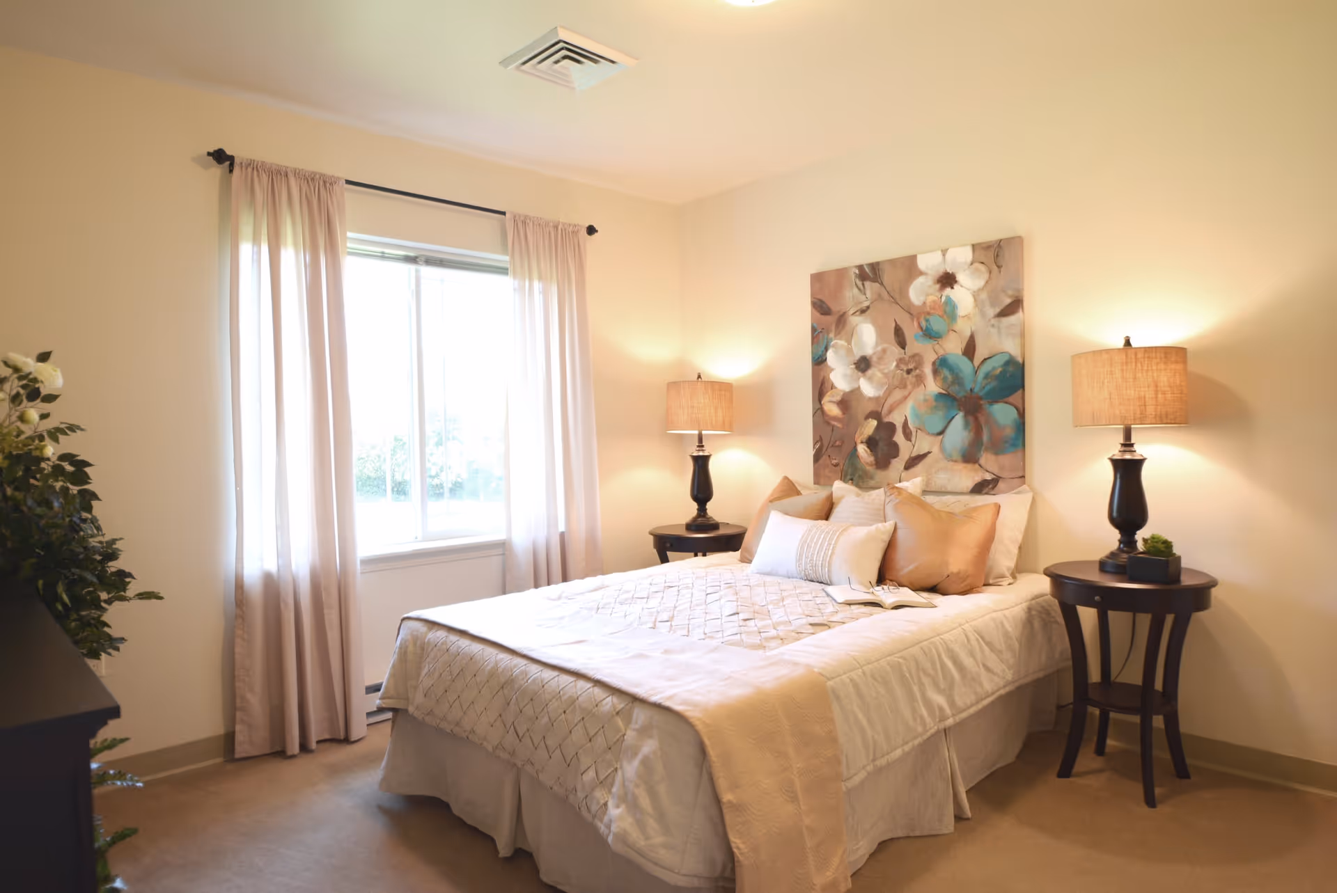 A cozy bedroom with a neatly made bed featuring beige and white bedding and multiple pillows. There are two dark wooden nightstands on either side of the bed, each with a lamp. A large floral painting with blue and white flowers hangs above the bed. A window with light beige curtains allows natural light into the room. A green plant is partially visible on the left side.