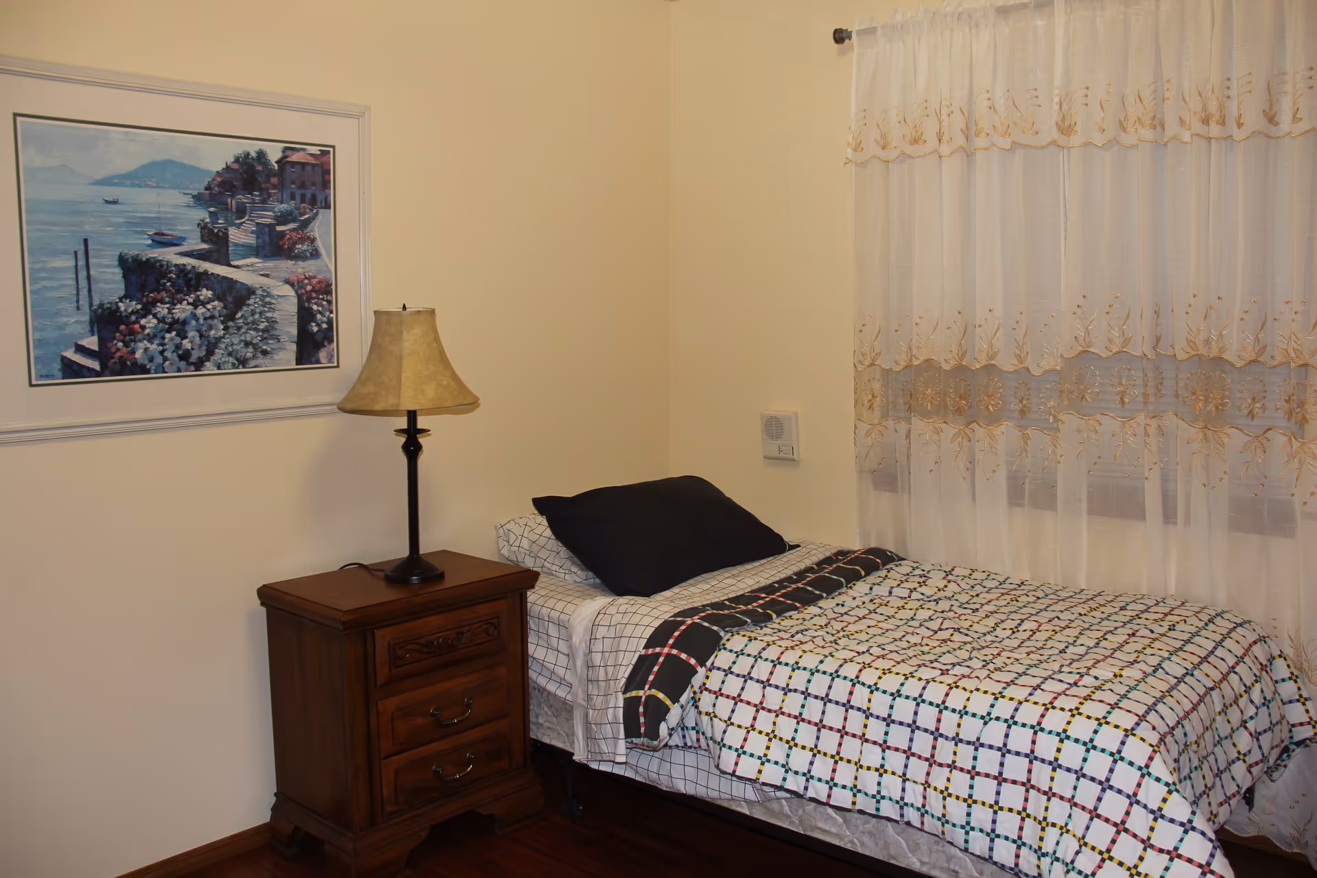 A small bedroom with a single bed covered in a colorful checkered quilt and a black pillow. Next to the bed is a wooden nightstand with two drawers and a table lamp. A framed painting of a coastal scene hangs on the wall above the nightstand. Sheer white curtains with gold embroidery cover the window.