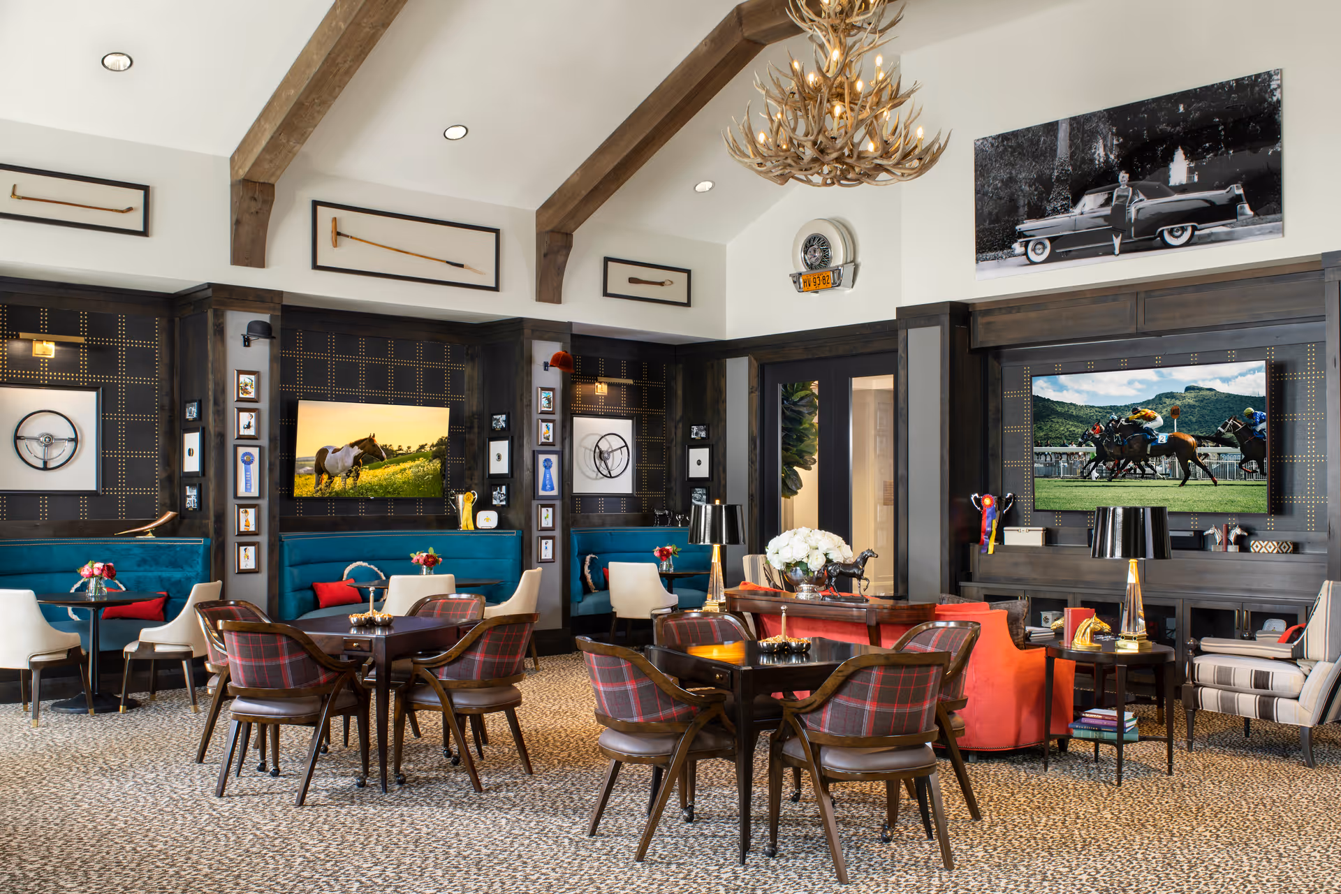A stylish and cozy common area with high vaulted ceilings and exposed wooden beams. The room features dark wood paneling, teal upholstered booth seating with red accent pillows, and several wooden tables with plaid cushioned chairs. The walls are decorated with framed pictures, vintage car steering wheels, and mounted antique golf clubs. Two large flat-screen TVs display images of a horse in a field and a horse race. The room is warmly lit with table lamps and wall sconces, and a large antler chandelier hangs from the ceiling.