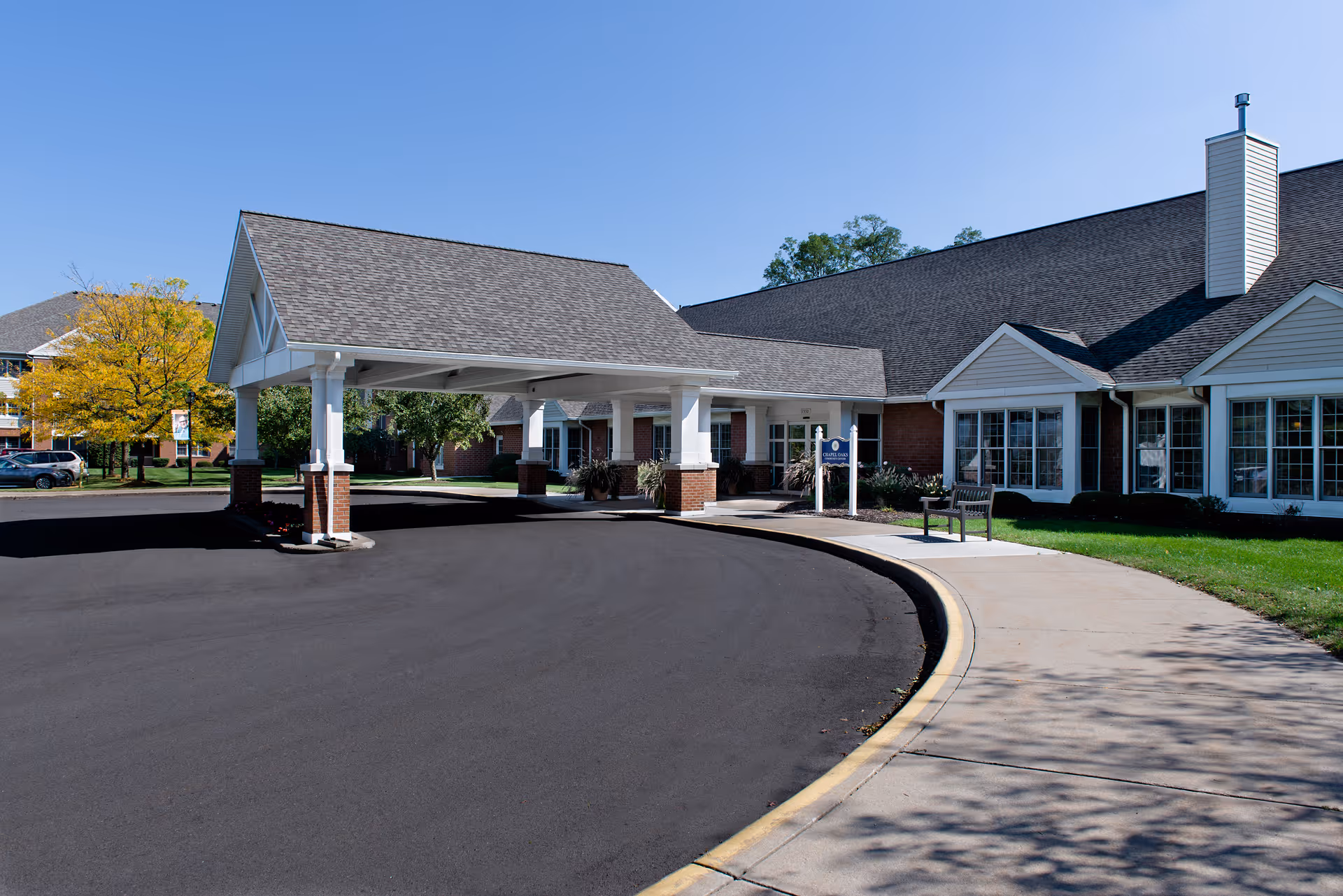 Exterior view of a senior living facility named Chapel Oaks featuring a covered entrance with white pillars and a curved driveway. The building has a brick and white siding facade with multiple windows and a chimney. There is a bench on the sidewalk and trees with green and yellow leaves in the background under a clear blue sky.