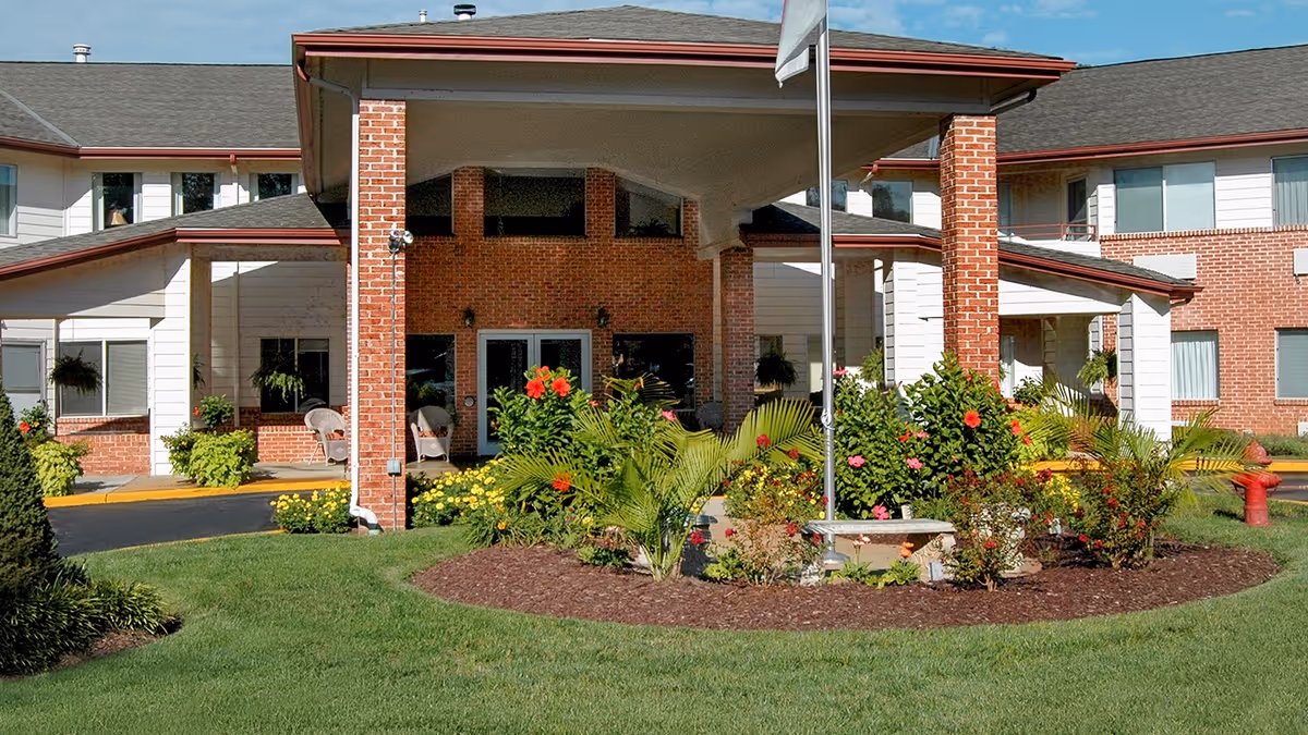 Front exterior view of Holiday Briarcrest Estates featuring a covered entrance with brick pillars, a landscaped garden with colorful flowers and shrubs, and a flagpole in front of the building.