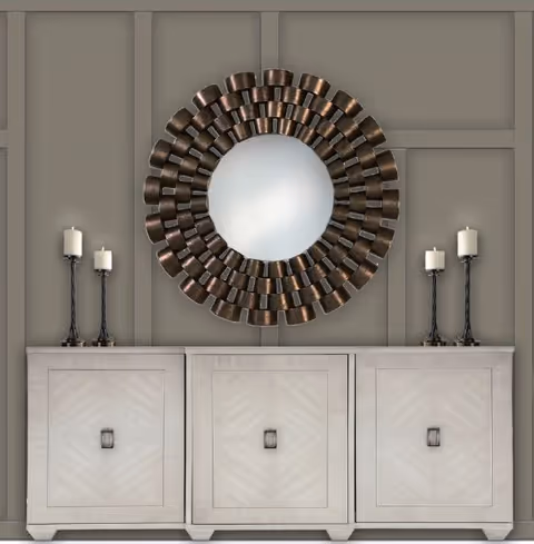A decorative interior wall featuring a large round mirror with a textured, dark metallic frame resembling overlapping cups. Below the mirror is a light-colored wooden cabinet with three sections, each with a square handle. On top of the cabinet are four tall candle holders, two on each side, each holding a white candle. The wall behind has a paneled design painted in a muted taupe color.