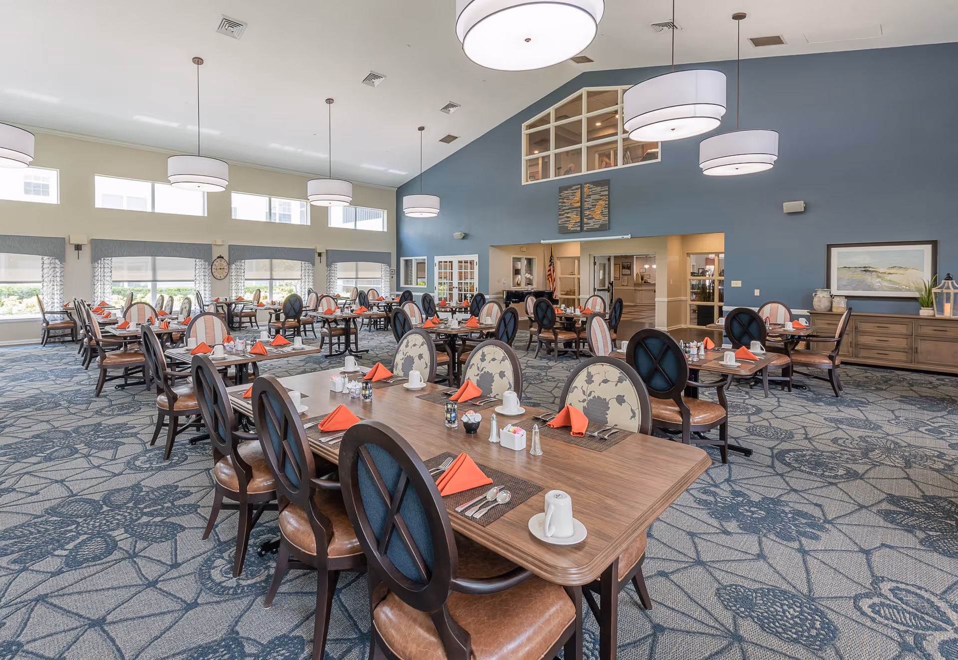 Spacious dining room with multiple tables set with white cups, silverware, salt and pepper shakers, and orange folded napkins. The room features large windows allowing natural light, patterned carpet, and modern hanging light fixtures. The walls are painted blue and beige with decorative artwork and a sideboard along one wall.