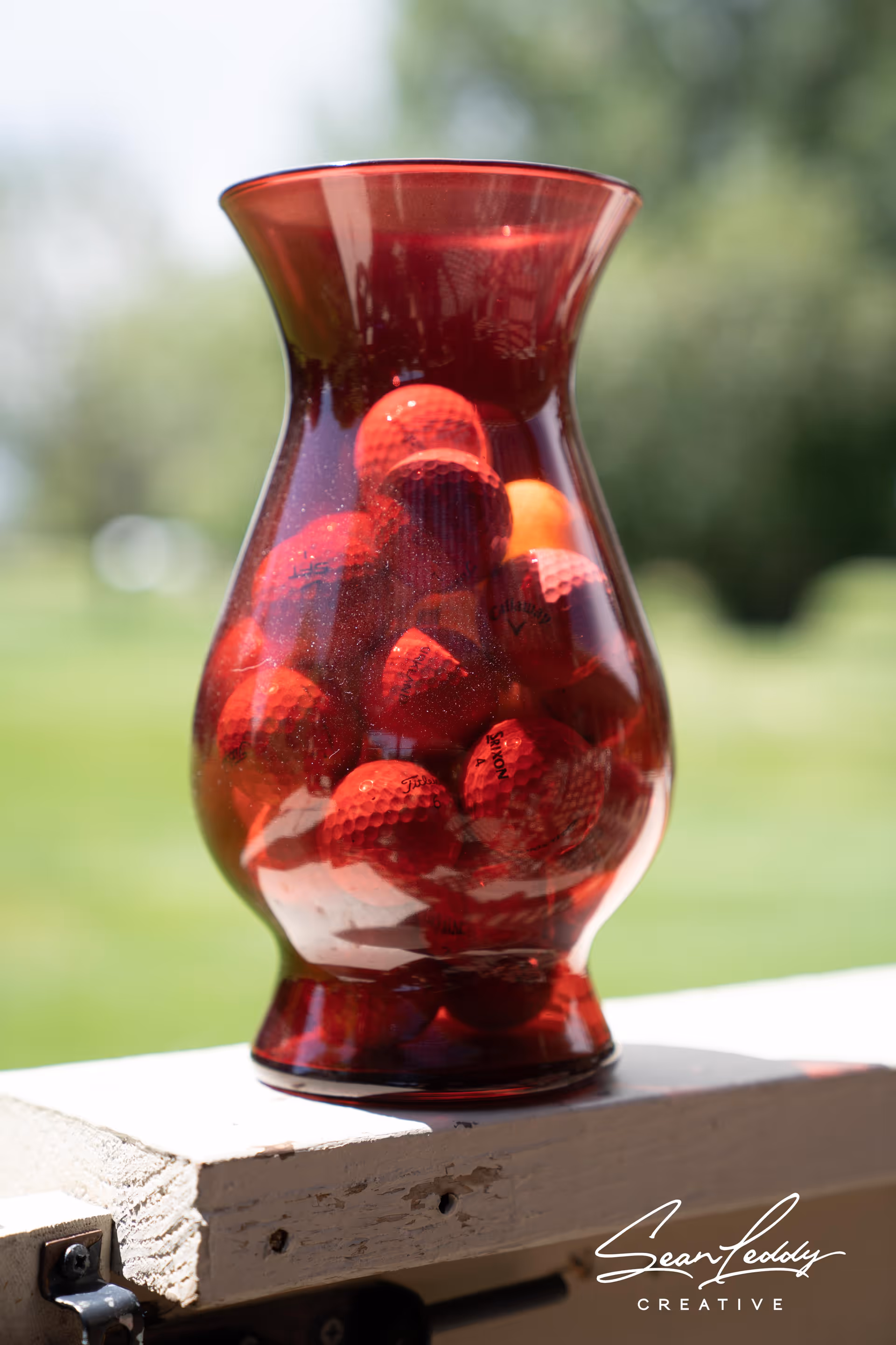 A red glass vase filled with red golf balls sits on a white wooden railing with a blurred green outdoor background.