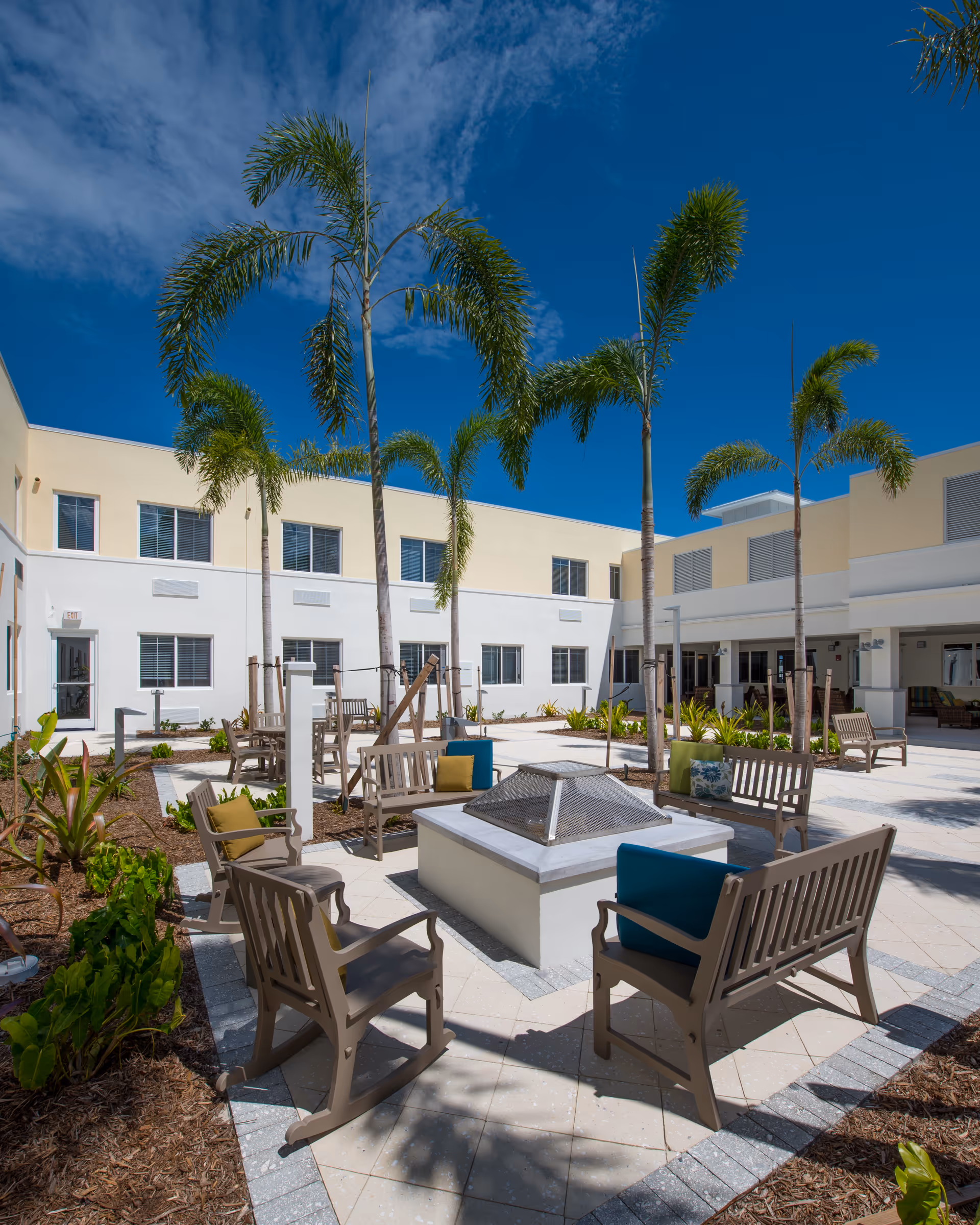 Sunlit courtyard with palm trees and multiple benches and chairs arranged around a central fire pit in front of a two-story residential building.