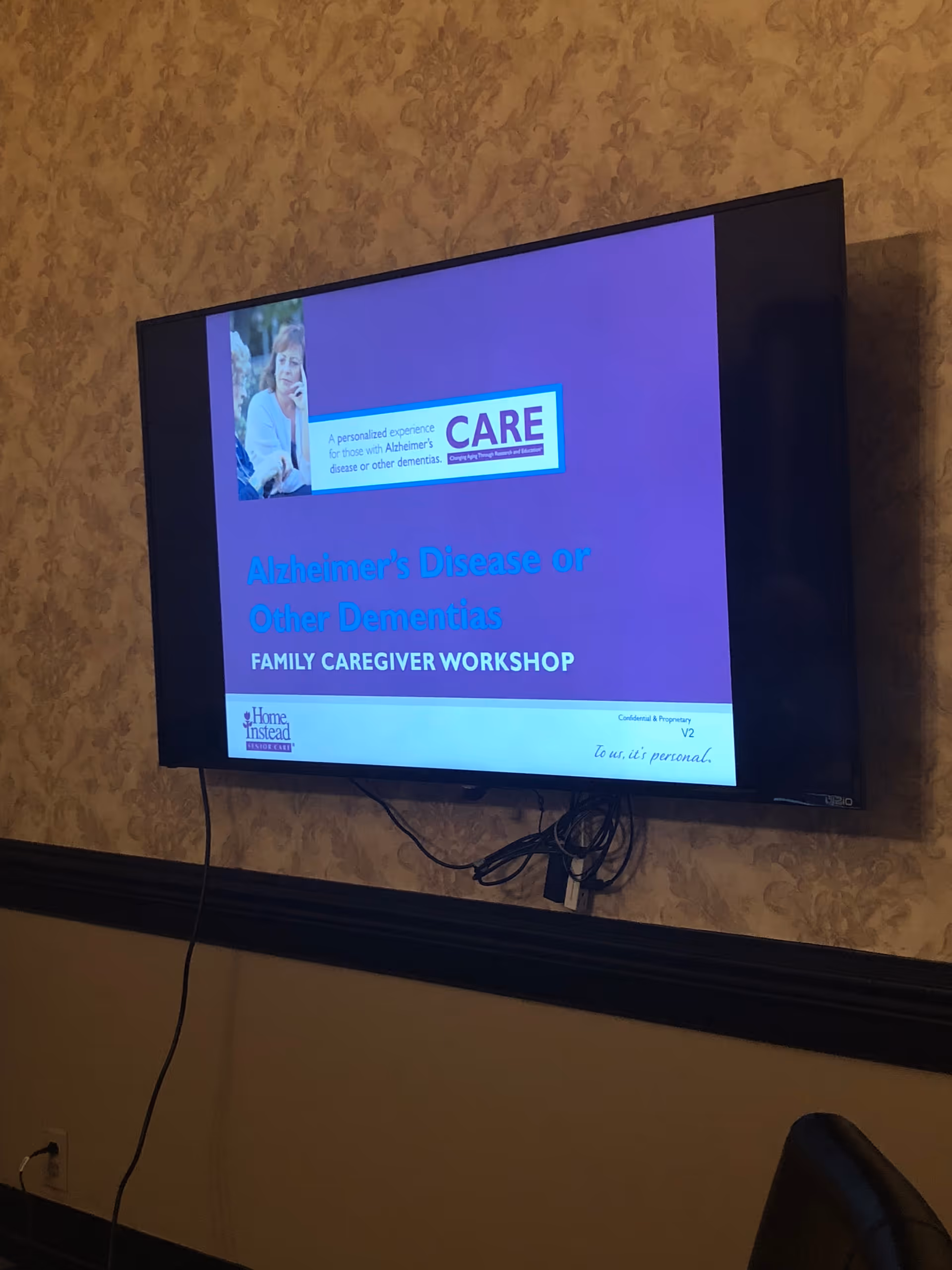 A wall-mounted flat screen TV displaying a presentation slide titled 'Alzheimer's Disease or Other Dementias FAMILY CAREGIVER WORKSHOP' with the Home Instead logo and a photo of an elderly woman. The TV is mounted on a beige patterned wallpapered wall with visible cables hanging below it.