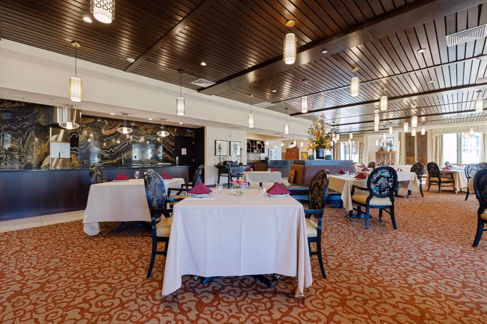 A spacious dining room with tables covered in white tablecloths, each set with folded burgundy napkins, glassware, and cutlery. The room features patterned carpet flooring, upholstered chairs with floral designs, and a wooden ceiling with multiple hanging pendant lights. A decorative floral arrangement is placed on a central divider, and large windows allow natural light to fill the space.
