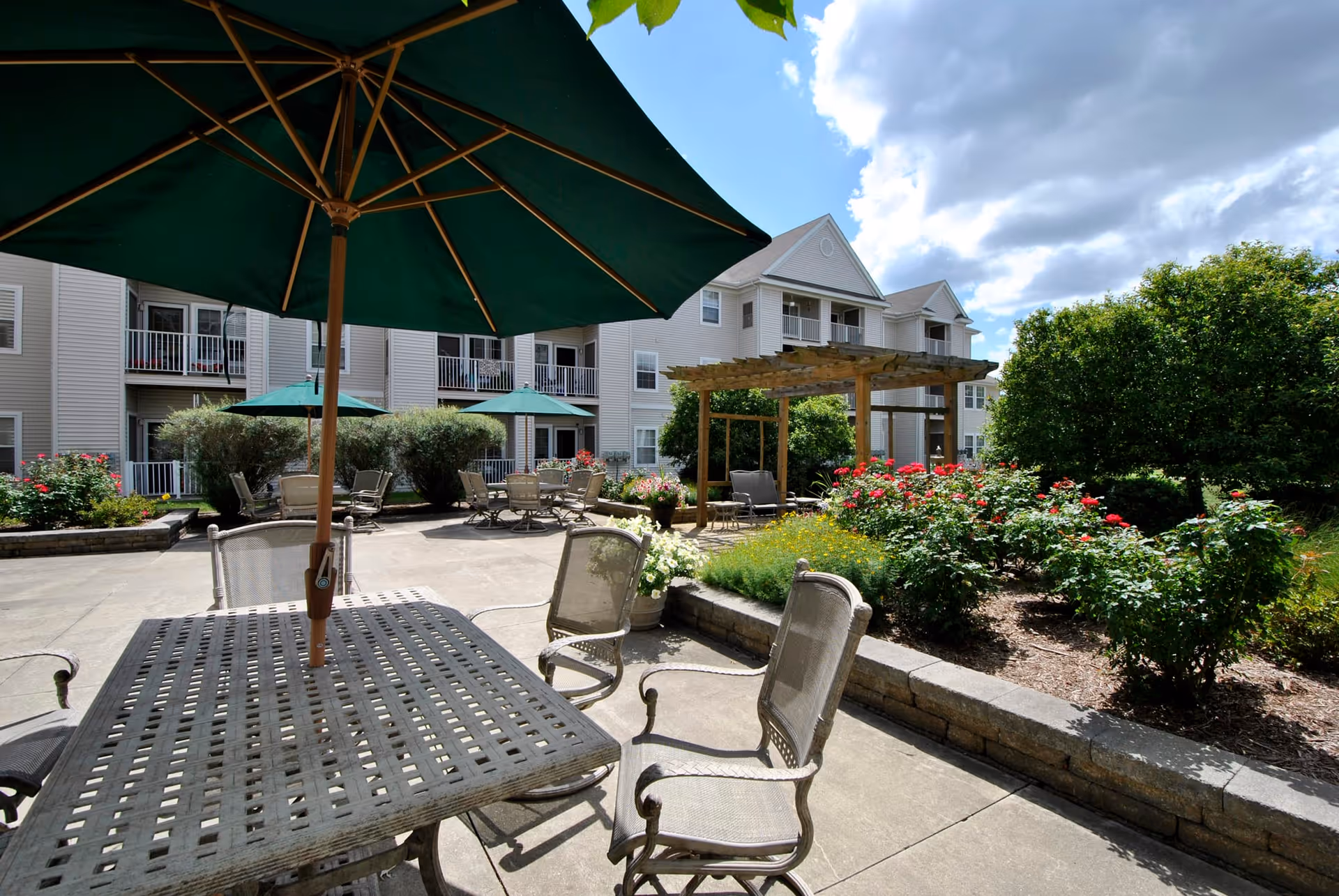 Outdoor patio area at Grandhaven Manor with metal tables and chairs, green umbrellas providing shade, a wooden pergola, and landscaped garden beds with flowering plants. The background shows a multi-story residential building under a partly cloudy sky.