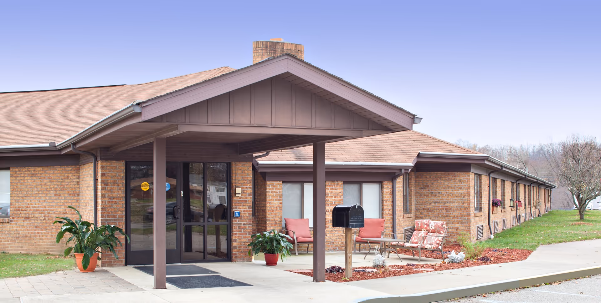Exterior view of a single-story brick building with a covered entrance supported by two columns. There are two potted plants near the entrance and outdoor seating with cushioned chairs and a small table on a landscaped area with mulch. The sky is clear and the surrounding area has grass and a few trees.