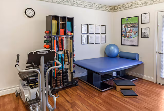 A small exercise room with wooden flooring featuring a stationary exercise bike, a rack holding various exercise bands and dumbbells, a large blue exercise mat with a blue exercise ball and a cylindrical bolster, and framed certificates and a wellness poster on the walls.