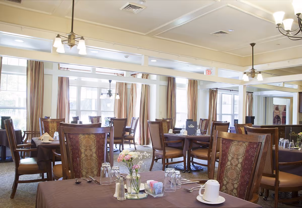 A bright dining room with multiple wooden tables and chairs. Tables are set with glasses, utensils, napkins, and small flower arrangements. Large windows with beige curtains allow natural light to fill the room. Ceiling lights and fans hang from the ceiling.