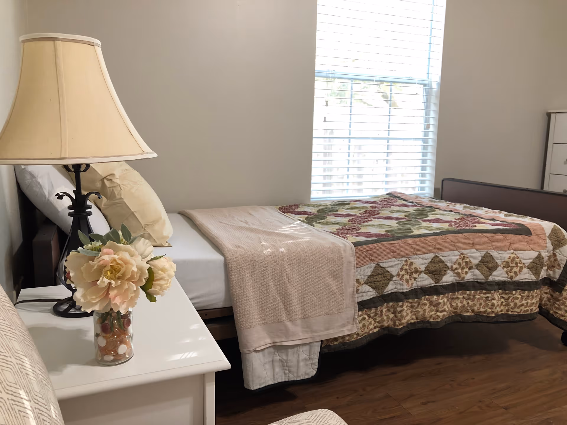A cozy bedroom with a single bed covered by a floral quilt and a beige blanket. Next to the bed is a white nightstand with a decorative lamp and a vase of flowers. A window with white blinds lets in natural light, and there is a white dresser in the corner.