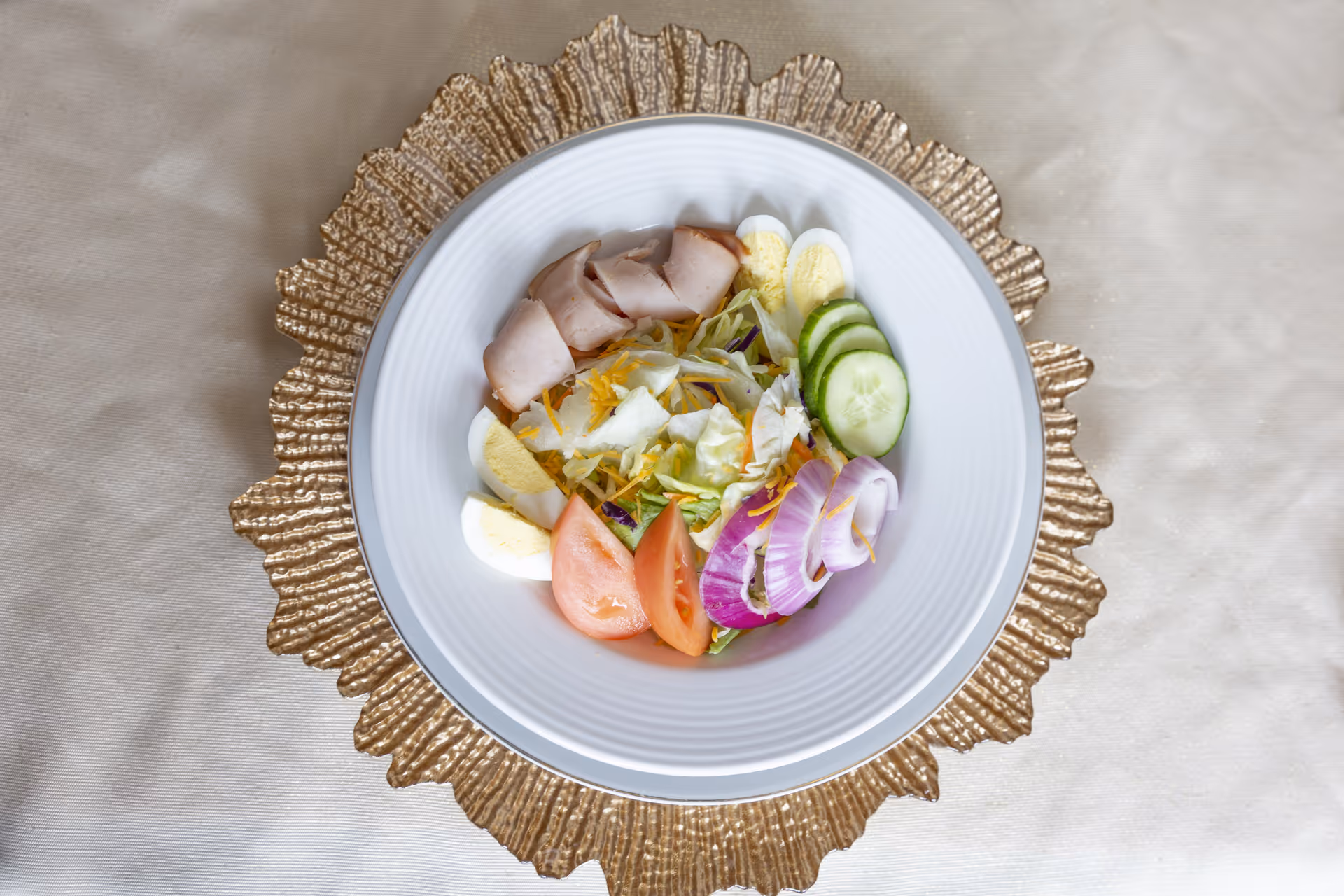 A white bowl filled with a fresh salad containing sliced turkey, hard-boiled egg halves, cucumber slices, tomato wedges, red onion rings, shredded cheese, and lettuce, placed on a decorative gold placemat on a beige tablecloth.
