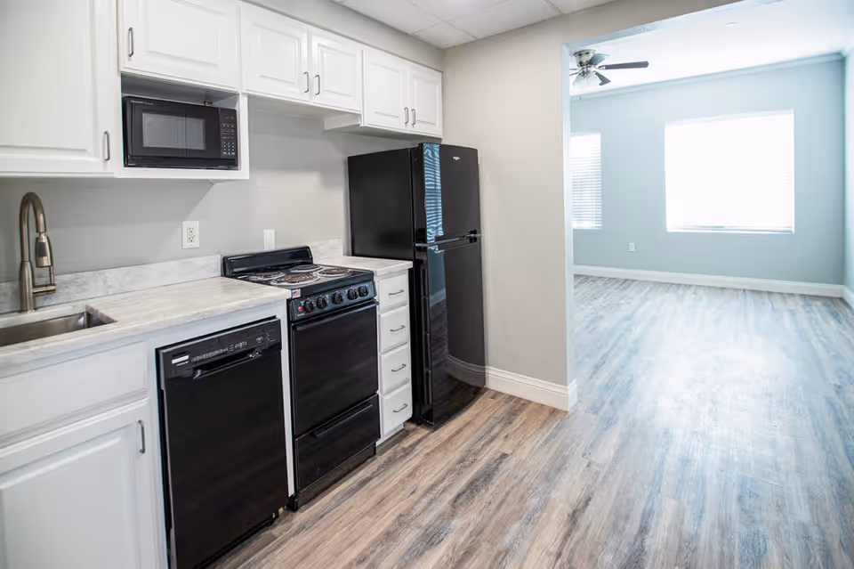 A modern kitchen area featuring white cabinets, a black microwave, black dishwasher, black stove, and black refrigerator. The kitchen has a light-colored countertop and a stainless steel sink with a faucet. The floor is covered with wood-style vinyl flooring that extends into an adjacent room with light blue walls and a ceiling fan with light.