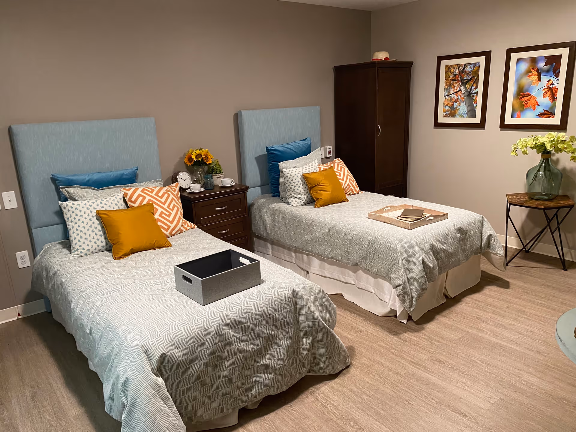 A cozy bedroom with two twin beds, each with light blue headboards and patterned bedding. The beds have multiple decorative pillows in blue, white, orange, and mustard yellow. Between the beds is a dark wooden nightstand with a clock, a vase of sunflowers, and a cup. On the right side of the room is a dark wooden wardrobe and a small table with a large glass vase containing green foliage. Two framed pictures of autumn leaves hang on the wall above the table. The floor is light wood, and the walls are painted beige.