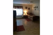 Small assisted living room with a couch/daybed, TV on a dresser, a side table lamp, a window with a valance, and framed photos on the wall.