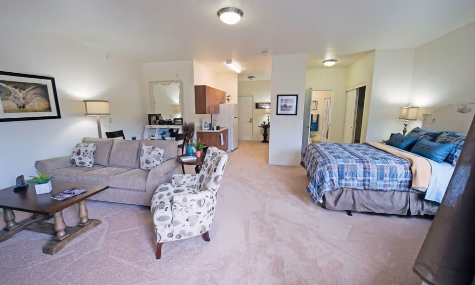 A spacious studio apartment featuring a living area with a beige sofa, patterned armchair, and wooden coffee table. The sleeping area has a bed with blue and beige plaid bedding. The room includes a small kitchen with wooden cabinets, a refrigerator, and a countertop. The walls are decorated with framed pictures and a large mirror. The carpet is light beige, and the room is well-lit with ceiling lights and lamps.