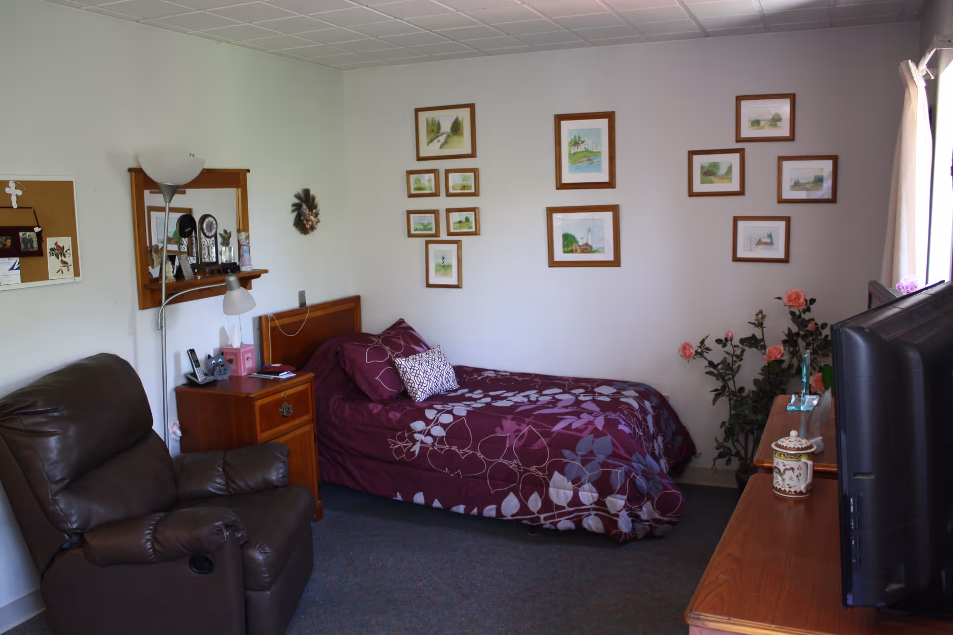 A cozy assisted-living bedroom with a single bed, reclining chair, TV, and framed artwork on the walls.