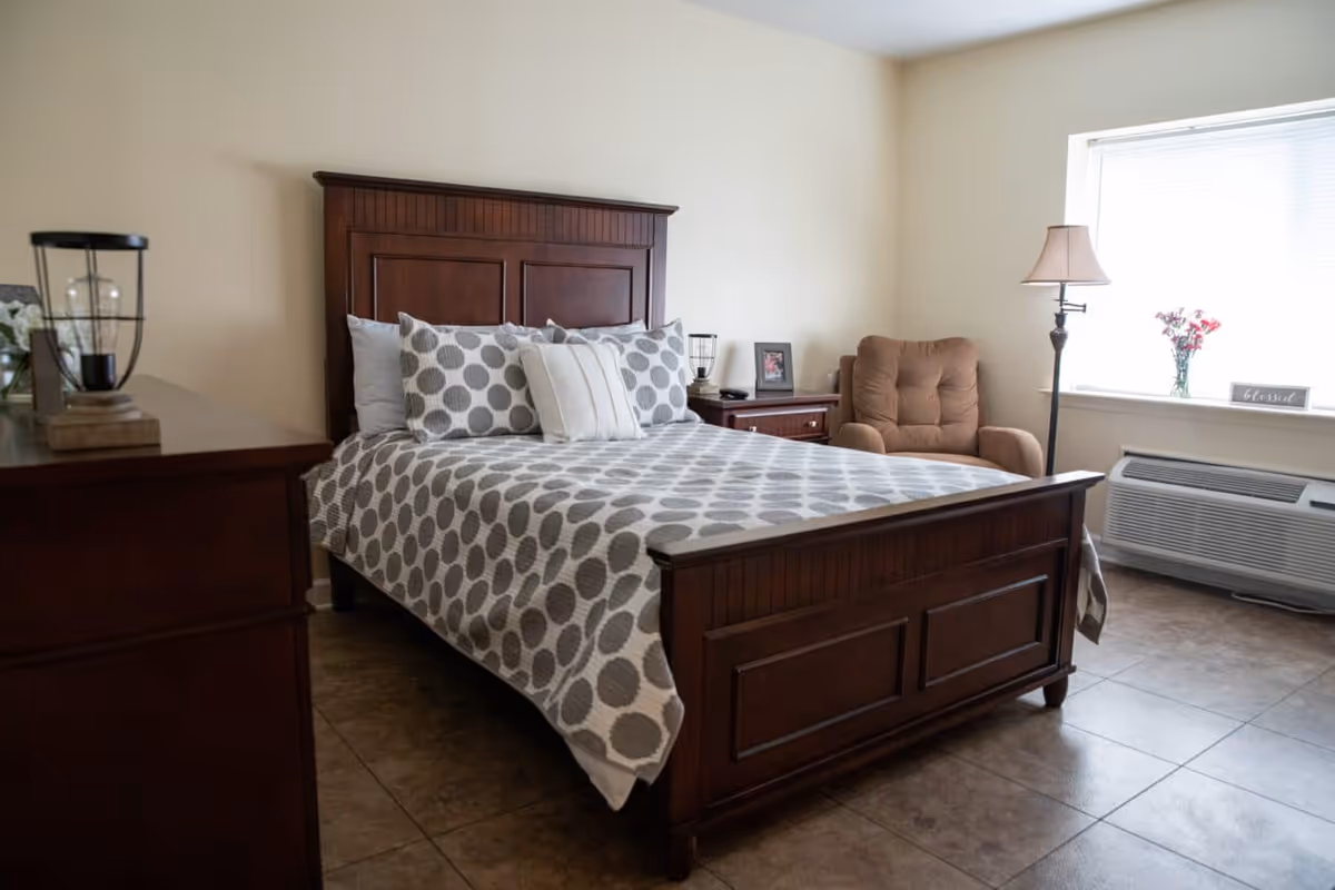A furnished bedroom with a wooden bed dressed in gray polka-dot bedding, a bedside table, armchair, floor lamp, and a window with an air conditioner.