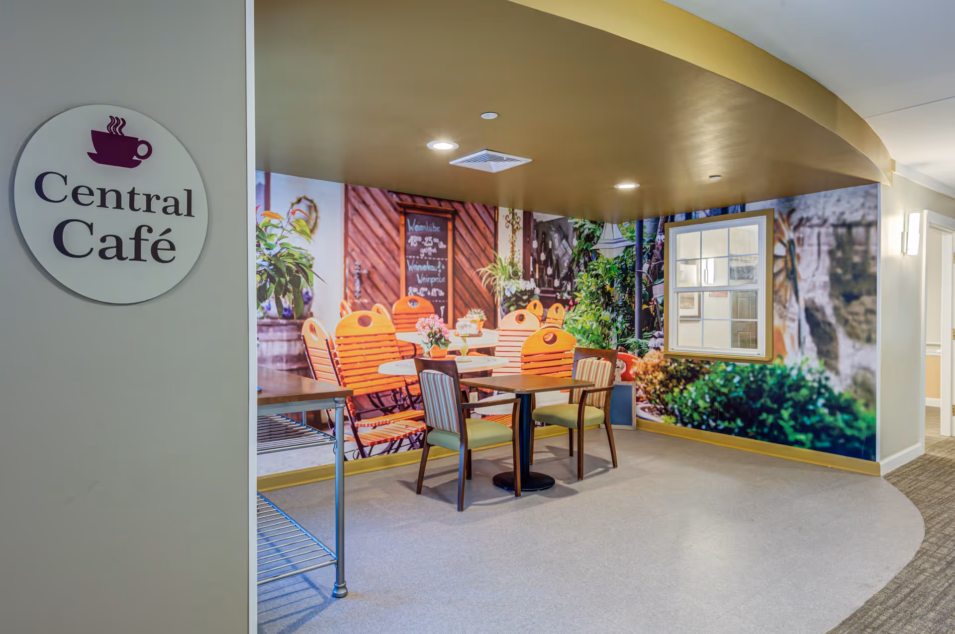 Interior view of a small café area named Central Café with a round sign on the wall featuring a steaming coffee cup icon. The café has a table with four chairs, two with striped upholstery and two with solid green seats. The back wall is decorated with a large mural depicting an outdoor garden scene with wooden chairs, plants, and a window, creating a cozy and inviting atmosphere.
