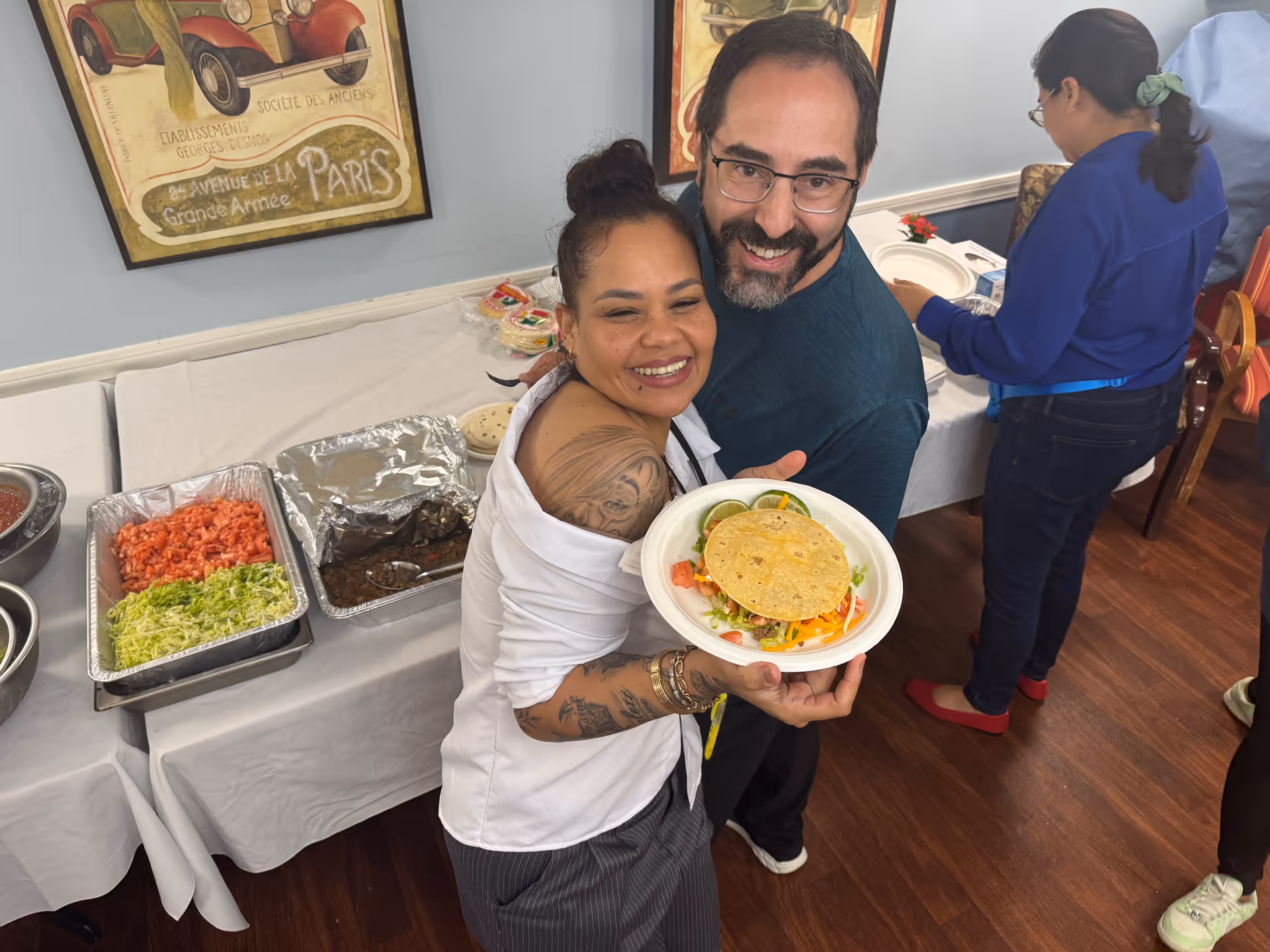 Two smiling adults holding a plate with tacos in front of a buffet table inside a dining room.