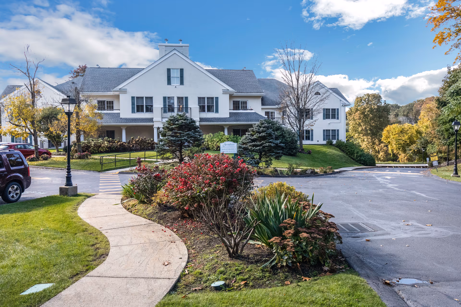Front exterior of a white two-story senior living building with a curved walkway, landscaped gardens, and a driveway.