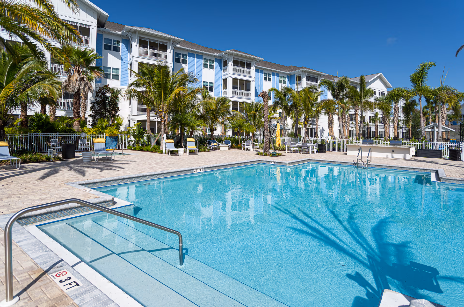 Outdoor swimming pool with clear blue water surrounded by palm trees and lounge chairs, with a multi-story residential building in the background under a clear blue sky.