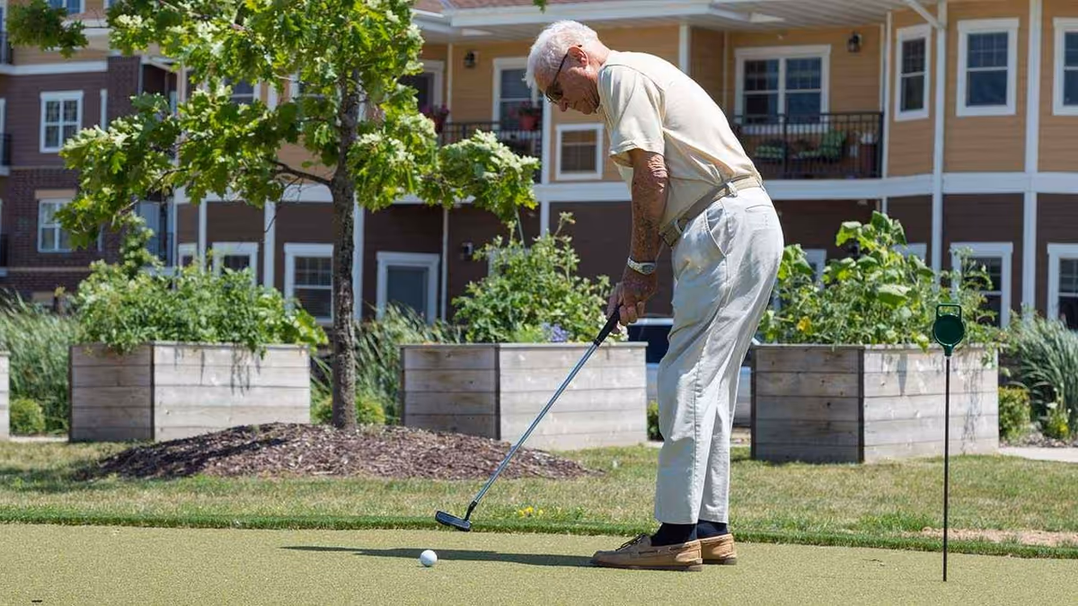 An elderly man playing mini golf on a putting green outside at Oak Park Senior Living. He is focused on putting the golf ball, with raised garden beds and a multi-story residential building in the background.