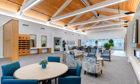 A spacious senior living common area with high wooden ceilings and exposed beams. The room features multiple seating arrangements including round tables with blue chairs, armchairs with floral patterns, and other cushioned chairs. Large windows allow natural light to fill the space, and there are plants placed throughout the room. A fireplace is visible on the right side of the image.