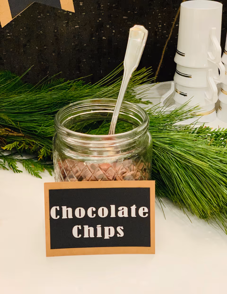 Glass jar with a spoon and a card labeled 'Chocolate Chips' set on a table with greenery and stacked cups.